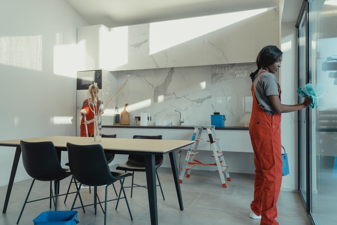 Two women in work clothes cleaning a kitchen with glass doors.